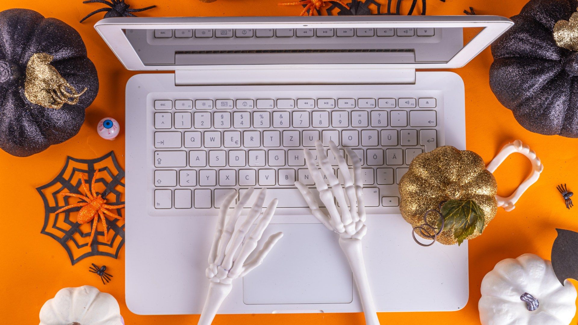 Computer portatile circondato da zucche, ragni e mani di scheletro su sfondo arancione: il contenuto si costruisce prima che la festa arrivi